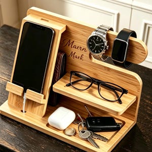 Peut inclure: Un organisateur de bureau en bois de bambou clair comprenant un support vertical pour smartphone avec câble de charge, une étagère pour lunettes et un plateau de base. Le bois est gravé "Marrn Mark" et contient une montre, une montre connectée, un boîtier d'écouteurs blanc, une clé de voiture et un trousseau de clés.