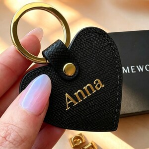 Op de afbeelding: Een zwarte hartvormige sleutelhanger met een textuur en een goudkleurige metalen ring. De naam Anna is in gouden letters op de voorzijde gedrukt. Een kleine goudkleurige metalen stud bevestigt het hart aan de ringbevestiging.