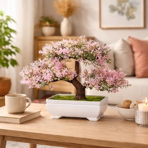 Peut inclure: Un bonsaï décoratif avec des fleurs roses et un feuillage vert est présenté dans un pot carré blanc. L'arbre a un tronc et des branches bruns. Le pot est sur une table en bois, avec une tasse et un livre à proximité.