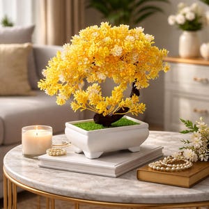 Peut inclure: Un bonsaï décoratif avec des fleurs jaune vif et un tronc brun foncé est placé dans un pot rectangulaire blanc rempli de mousse verte. Le bonsaï est sur une table en marbre avec une bougie allumée et des bijoux.