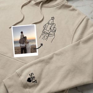 Könnte beinhalten: Beiger Kapuzenpullover mit Kordelzug und Vordertasche. Mit einem schwarzen, gestickten Familienporträt und den Initialen "S&F" mit einem Herz auf dem Ärmel. Enthält ein Foto einer Familie am Strand.