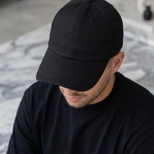 Könnte beinhalten: Schwarze Baseballkappe mit der römischen Zahl "LII MMXXIV" auf der Vorderseite gestickt. Die Kappe hat einen gebogenen Schirm und eine strukturierte Passform. Das Bild zeigt die Kappe aus einem leicht erhöhten Winkel.