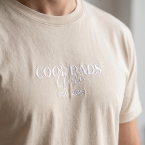 Könnte beinhalten: Beiges T-Shirt mit weiß gesticktem Schriftzug "COOK DADS Club est. 2026". Das Shirt hat einen Rundhalsausschnitt und kurze Ärmel. Der Stoff wirkt weich und leicht strukturiert.