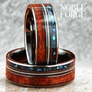 May include: Two rings with a wooden band and a silver metal inlay. The rings have a dark brown wood grain and a band of iridescent blue and green pieces. The text "NOBLE BANDS FORGE" is in the background.