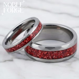 Puede incluir: Dos anillos plateados con textura martillada y una incrustación roja vibrante. Los anillos son probablemente alianzas de boda, que muestran un diseño moderno con un contraste de color audaz. Los anillos se exhiben sobre una superficie de mármol.