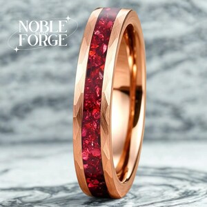 May include: Rose gold ring with a faceted band and a vibrant red gemstone inlay. The ring features the text "NOBLE FORGE" on the left side. The ring's design is modern and eye-catching, suitable for everyday wear or special occasions.