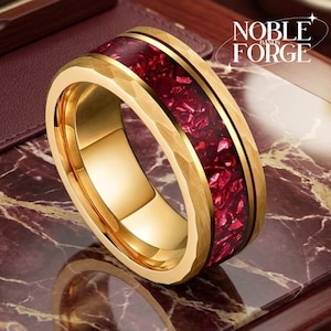 May include: A gold-toned ring featuring a hammered finish on the outer edges and a central inlay of crushed deep red material. The text Noble Forge is visible in the upper right corner of the image, accompanied by a small four-pointed star graphic.