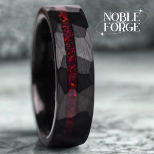 May include: A black ring with a faceted surface and a vibrant red inlay. The ring has a modern design, with the text "NOBLE FORGE BANDS" in white on the right side. The ring is a bold statement piece.