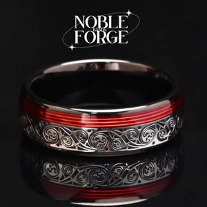 Puede incluir: Un anillo plateado con una franja roja y un intrincado trabajo de volutas. El anillo tiene un interior negro y las palabras "NOBLE FORGE" se muestran en escritura blanca.