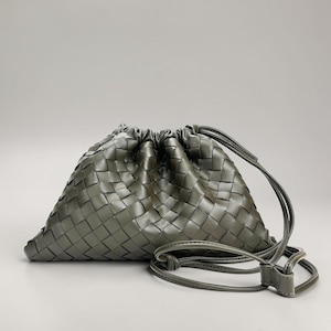May include: A woven, olive-green drawstring bag with a long, thin strap. The bag's body is a triangular shape, with a gathered top. The strap is attached to the bag with a small, rectangular piece.