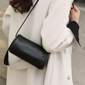 Puede incluir: Un bolso bandolera negro, cilíndrico, con una correa larga y fina. El bolso está hecho de un material texturizado y presenta una pequeña hebilla rectangular. La correa es negra y el bolso se lleva sobre un abrigo blanco.