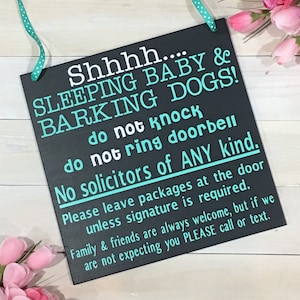 May include: A black rectangular sign with white and teal text, including "Shhhh... SLEEPING BABY & BARKING DOGS!" and "No solicitors of ANY kind." A teal and white polka dot ribbon is attached for hanging. Pink flowers are in the background.