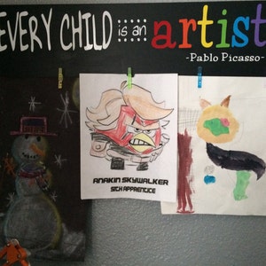 May include: Three children's drawings are displayed on a chalkboard with the quote "Every child is an artist -Pablo Picasso-." The drawings include a snowman, a red-haired character with a black jacket, and a green and yellow creature.