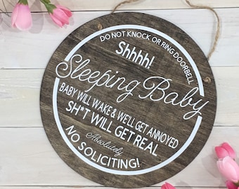 May include: A round wooden sign with a white border and white text that reads "DO NOT KNOCK OR RING DOORBELL Shhhh! Sleeping Baby BABY WILL WAKE & WE'LL GET ANNOYED SH*T WILL GET REAL Absolutely NO SOLICITING!"