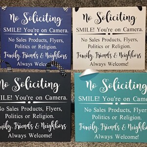 May include: Four wooden signs with the text "No Soliciting" in white lettering. Each sign has a different background colour: blue, white, grey, and teal. The signs all have the same message: "No Soliciting, SMILE! You're on Camera. No Sales Products, Flyers, Politics or Religion. Family, Friends & Neighbours Always Welcome!"