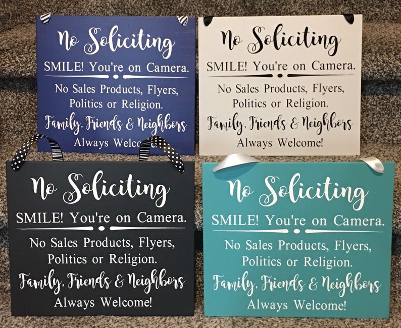May include: Four wooden signs with the text "No Soliciting" in white lettering. Each sign has a different background colour: blue, white, grey, and teal. The signs all have the same message: "No Soliciting, SMILE! You're on Camera. No Sales Products, Flyers, Politics or Religion. Family, Friends & Neighbours Always Welcome!"