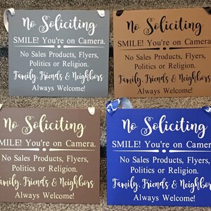 May include: Four wooden signs with the text "No Soliciting" in white lettering. Each sign has a different background colour: grey, brown, brown, and blue. The text on each sign reads: "SMILE! You're on Camera. No Sales Products, Flyers, Politics or Religion. Family. Friends & Neighbours Always Welcome!"