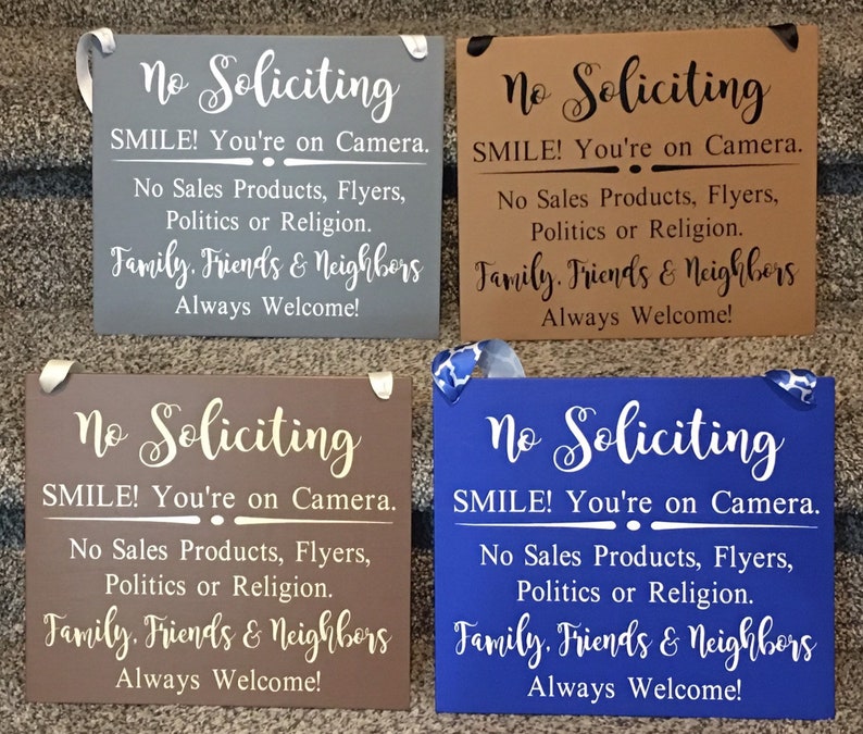 May include: Four wooden signs with the text "No Soliciting" in white lettering. Each sign has a different background colour: grey, brown, brown, and blue. The text on each sign reads: "SMILE! You're on Camera. No Sales Products, Flyers, Politics or Religion. Family. Friends & Neighbours Always Welcome!"