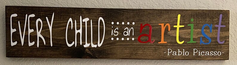 May include: A wooden sign with the text "EVERY CHILD is an artist - Pablo Picasso -" in white lettering. The word "artist" is spelled out in rainbow colors.