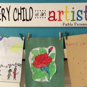 May include: A turquoise wall with a white sign that reads "EVERY CHILD is an artist -Pablo Picasso-".  There are three pieces of children's artwork hanging on the wall with clothespins. The artwork includes a drawing of three stick figures, a red flower, and a card that says "Mommy I like you" with red dots.