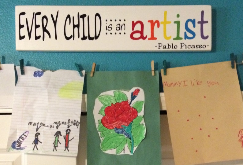 May include: A turquoise wall with a white sign that reads "EVERY CHILD is an artist -Pablo Picasso-".  There are three pieces of children's artwork hanging on the wall with clothespins. The artwork includes a drawing of three stick figures, a red flower, and a card that says "Mommy I like you" with red dots.