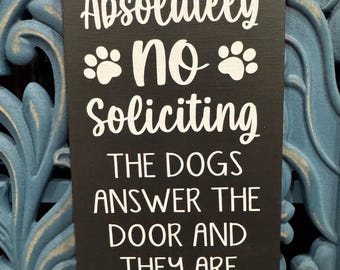 May include: A rectangular black hanging sign with white text that reads Absolutely no Soliciting, followed by two paw prints, and the text THE DOGS ANSWER THE DOOR AND THEY ARE ASSHOLES. The sign is suspended by a thin black and white string.
