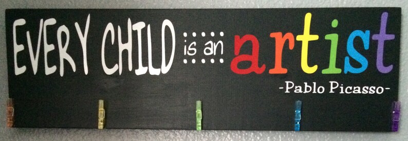 May include: Black chalkboard sign with the quote "EVERY CHILD is an artist - Pablo Picasso -" in white text. The word "artist" is written in rainbow colors. There are five colorful clothespins attached to the sign.