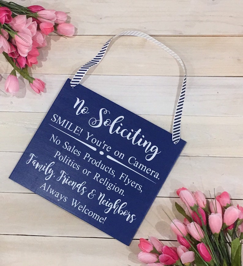 May include: A blue wooden sign with white text that reads "No Soliciting" and "SMILE! You're on Camera." The sign also says "No Sales Products, Flyers, Politics or Religion." and "Family, Friends & Neighbours Always Welcome!"