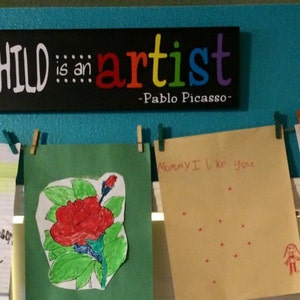 May include: A turquoise wall with a black sign that reads "Every child is an artist - Pablo Picasso".  There are several colorful drawings pinned to the wall with clothespins.