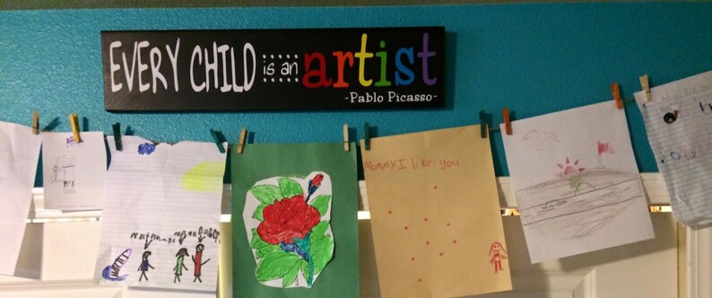 May include: A turquoise wall with a black sign that reads "Every child is an artist - Pablo Picasso".  There are several colorful drawings pinned to the wall with clothespins.