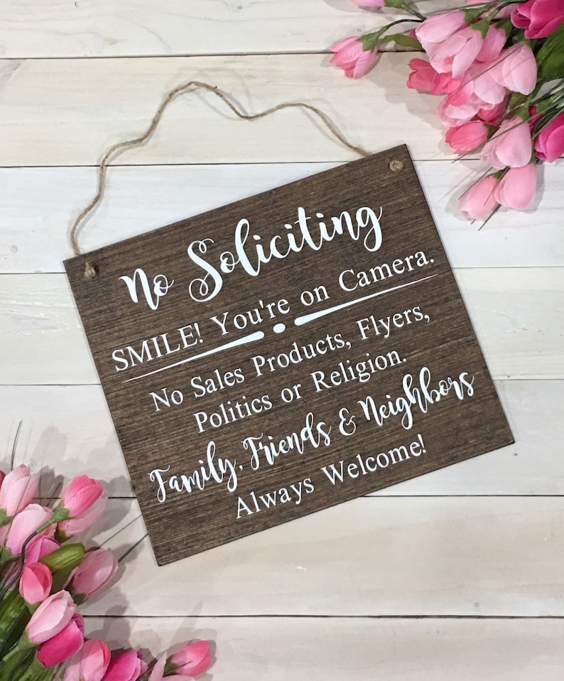 May include: A brown wooden sign with white text that reads "No Soliciting". The sign also includes the text "SMILE! You're on Camera. No Sales Products, Flyers, Politics or Religion. Family, Friends & Neighbours Always Welcome!"