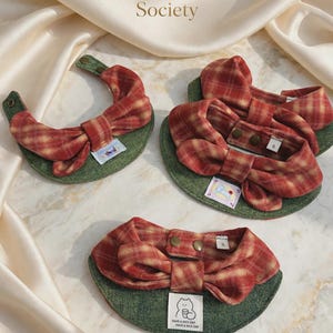 May include: Three pet bibs featuring a red and yellow plaid bow attached to a green textured base. Each bib has metal snap closures and a small white label. One label displays a line drawing of a cat with the text Have a nice day.
