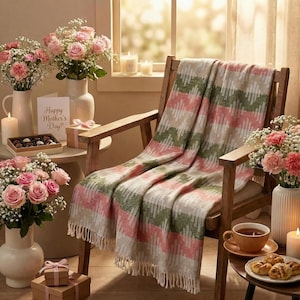 May include: A comfortable setting with a wooden chair and a blanket with a pink, green, and beige zigzag pattern. The room is decorated with pink roses and carnations in white vases, candles, and a small table with treats and a "Happy Mother's Day" card.
