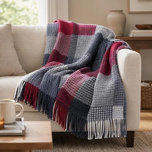 May include: A patterned throw blanket in red, grey, and navy blue, draped over a beige sofa. The blanket has a checkered design and fringed edges. A white mug is on a nearby table.