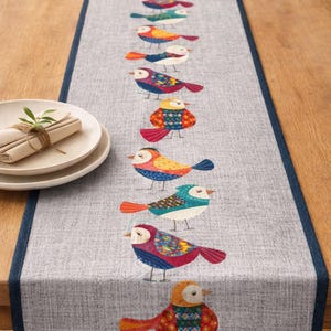 May include: A grey table runner decorated with a repeating pattern of vibrant, stylised birds. The birds display diverse patterns and colours, including red, blue, orange, and green. The runner has a dark blue border and is on a wooden table.