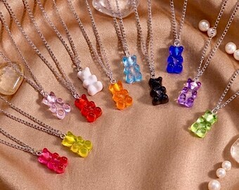 Könnte beinhalten: Eine Sammlung von Halsketten mit silbernen Ketten, die jeweils einen bunten Gummibären-Anhänger tragen. Die Anhänger sind in verschiedenen Farben erhältlich, darunter Rosa, Gelb, Grün, Lila, Schwarz, Blau, Orange und Weiß. Die Halsketten liegen auf einem beigen Stoff.