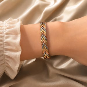 May include: A tri-color bracelet with a herringbone design. The bracelet features gold, silver, and rose gold tones. The jewelry is worn on a wrist, against a backdrop of a silky, neutral-toned fabric.