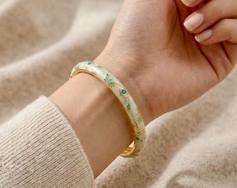 May include: A gold-toned bangle bracelet with a floral design. The bracelet features a pale green enamel base with blue and green floral accents. The bracelet is worn on a wrist, showcasing the intricate details of the jewelry.
