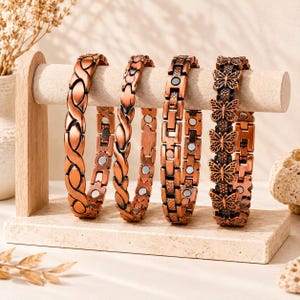 May include: Four copper-toned metal bracelets displayed on a stand. The first two feature an interlocking twisted design, the third has a rectangular link pattern with small circular accents, and the fourth displays a series of butterfly motifs.