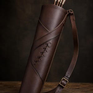 May include: A brown leather quiver with a shoulder strap, holding several arrows with white and brown fletching. The quiver has decorative stitching and a buckle closure. The leather has a rich, dark color, and the design suggests a traditional archery theme.