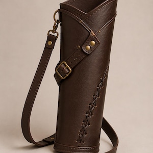 May include: A brown leather quiver with a shoulder strap and buckle closure. The quiver has decorative stitching down the side and brass-colored hardware. The design is cylindrical with a flared top.