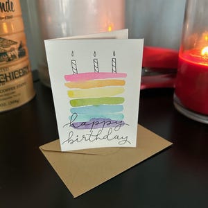 May include: A watercolor birthday card featuring a colorful cake design with three lit candles. The cake has layers of pink, yellow, green, blue, and purple. The words "happy birthday" are written in cursive below the cake. The card rests on a brown envelope.