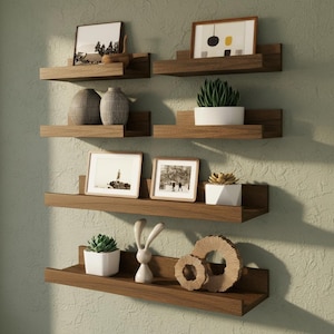 May include: A set of five wooden floating shelves mounted on a wall, each holding decorative items. The shelves display framed pictures, potted plants, and ceramic vases, creating a modern and organized display.