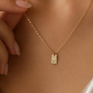Könnte beinhalten: Eine goldfarbene Halskette mit einer zarten Kette und einem kleinen, rechteckigen Anhänger. Der Anhänger ist mit dem Großbuchstaben M in einer Serifenschrift graviert. Die Kette ist dünn und besteht aus Gliedern, die eine einfache Schlaufe bilden.