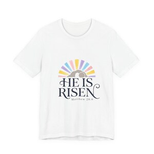 Può includere: T-shirt bianca con la scritta blu navy "HE IS RISEN" e un design colorato di una tomba e raggi di luce. Il testo "Matthew 28:6" è in basso.