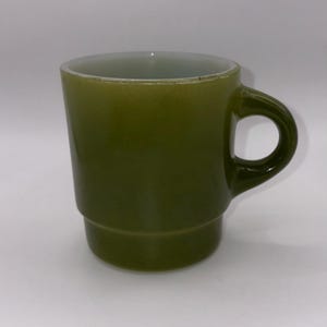 May include: A vintage green glass mug with a rounded handle. The mug has a slightly flared shape and a two-tiered design. The interior is a lighter shade of green. The mug is likely from the mid-20th century.