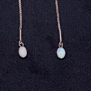 May include: A pair of silver-toned drop earrings. Each earring features a delicate chain suspending a small, oval, opalescent stone. The stones have a soft, iridescent glow against a dark background.