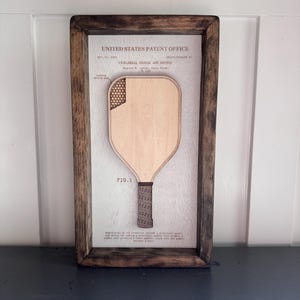 May include: A framed wall art piece featuring a United States Patent Office document for a pickleball paddle and method. The display includes a light wood cutout of a paddle with a honeycomb pattern in the corner, set against a white background.