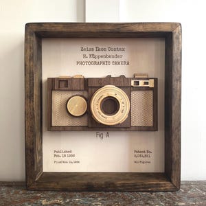 May include: Wooden framed artwork featuring a detailed, three-dimensional replica of a Zeiss Ikon Contax photographic camera. The artwork includes the text "Zeiss Ikon Contax" and "PHOTOGRAPHIC CAMERA". The frame is dark brown.