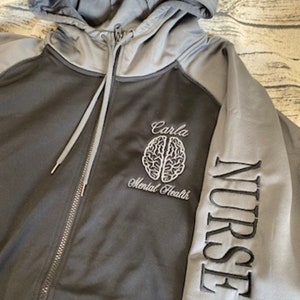 May include: A gray and black zip-up hoodie with the word "NURSE" embroidered on the right sleeve. The front of the hoodie features a white embroidered brain with the text "Carla Mental Health".